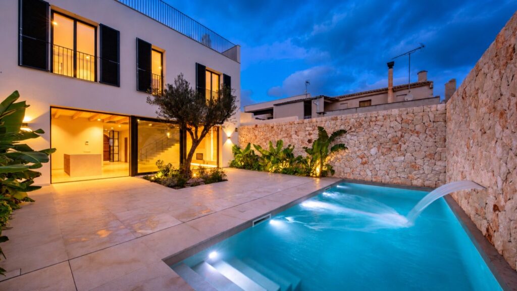 Patio trasero con piscina y cascada al anochecer en la casa adosada de Ses Salines, con muro de piedra y olivo